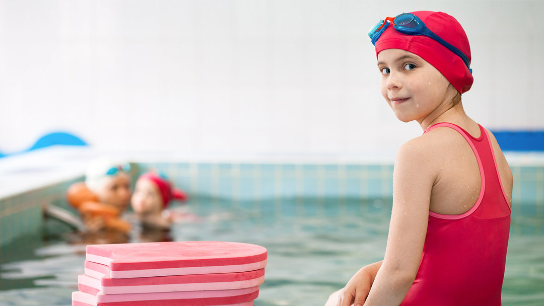 Girls sitting on the edge of the pool next to a pile of floats. Her back is to the camera and she is looking over her shoulder towards us.